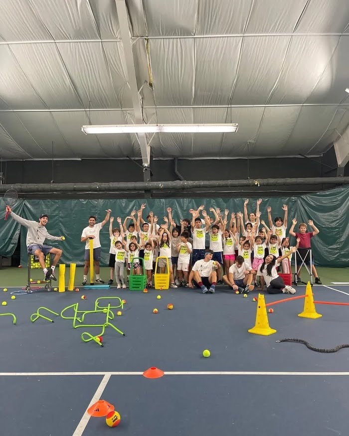 Children and coaches at a tennis academy celebrate after a fun indoor tennis camp session, surrounded by training cones, hurdles, tennis balls, and practice equipment on the court.